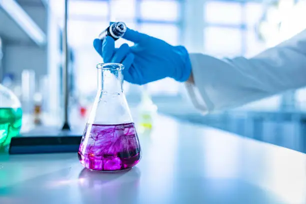Close up of unrecognizable chemist pouring purple liquid into a beaker at laboratory. Focus is on flask. Copy space.