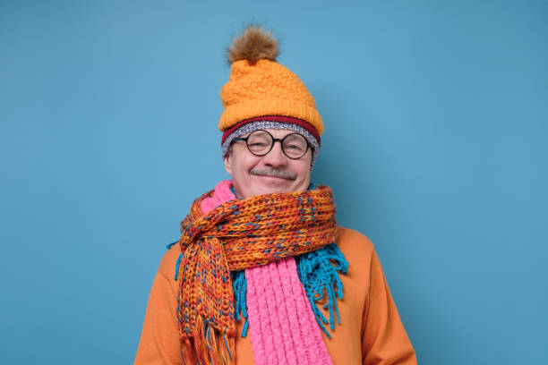Handsome caucasian funny senior man in several hats and scarfs on blue background. Winter fashion.