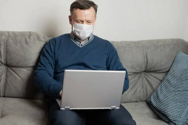 Photo of Adult man in protective medical mask work at home by laptop. Remote work. Covid-19. Quarantine. Stay at home.