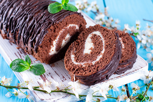 Delicious chocolate roll sponge cake with vanilla cream, mint leaves and spring flowers on a blue background. Desert sweet food