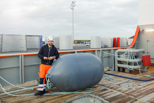 Technician standing on transfer vessel deck and leaning on a gigantic float and with parts of wind-turbines behind
