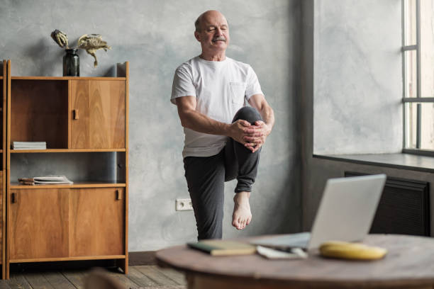 man doing yoga exercise at home using online lesson on notebook. Senior handsome sportman doing stretching and balancing yoga exercise at home using online lesson on notebook. How to stay healthy on quarantine concept. good posture stock pictures, royalty-free photos & images