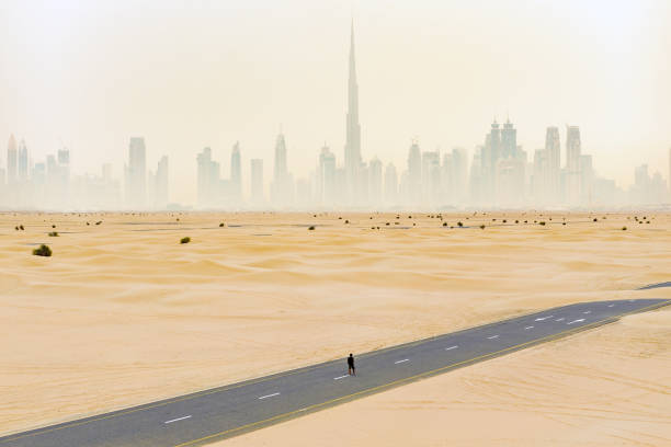 view from above, stunning aerial view of an unidentified person walking on a deserted road covered by sand dunes with the dubai skyline in the background. dubai, united arab emirates. - fog desert arabia sunset imagens e fotografias de stock