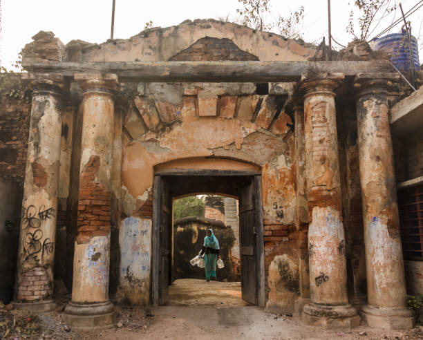 Murshidabad, West Bengal/India - January 15 2018: The old, crumbling ruins of an ancient stone gateway on the streets of Murshidabad. A woman stands behind the door holding water cans in her hand.