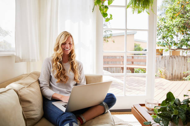 istock Smiling young blonde woman using a laptop on her sofa 1218020242