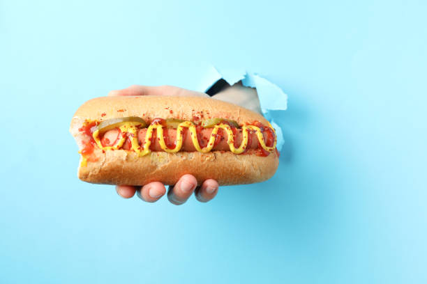 istock Hand holds hot dog from hole on blue background 1218020338