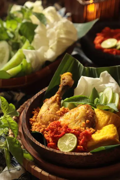Ayam Goreng Penyet, the popular East Javanese smashed, fried chicken dish. The fried chicken pieces are smashed with a pestle before being plated. It served together with red chili paste, tempeh, tofu, and fresh vegetables. The dish is plated on a wooden plate lined with banana leaf.