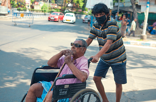 Glimpses of domestic life and city under lockdown facing threat of coronavirus epidemic in India. A disabled senior adult wearing black spectacle (either due to blindness or low vision) in a wheelchair being cared for by a middle aged person in street. One of them is wearing face mask except the aged person, who is at greater risk during coronavirus epidemic.\n\nAs of 10th April, 2020, confirmed cases in India is 6,761 while 206 people have died after being tested Covid-19 positive.\n\nPhoto taken in Kolkata on 04/09/2020, 16th day of pan India lockdown.