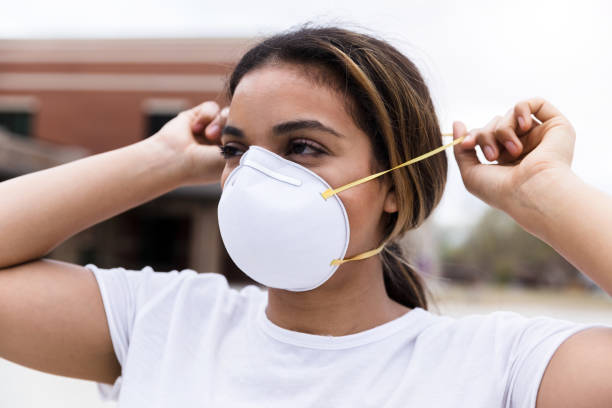 Mid adult woman putting on N95 face mask During a pandemic, a mid adult woman protects herself by placing an N95 face mask over her nose and mouth. She is standing outdoors. n95 face mask photos stock pictures, royalty-free photos & images
