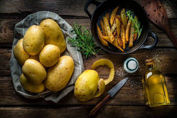 Preparing homemade french fries: top view of a rustic kitchen table with ingredients and kitchen utensils for preparing french fries ah home. A raw potatoes heap is at the left arranged on a fabric tissue. A peeled potato and an old kitchen knife are beside the heap. A cast iron cooking pan with cooked french fries is at the top right and a salt container and cooking oil bottle are at the bottom right.  Some rosemary branches complete the composition. Predominant colors are yellow and brown. High resolution 42Mp studio digital capture taken with SONY A7rII and Zeiss Batis 40mm F2.0 CF lens