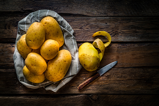 Raw potatoes heap shot from above on rustic wooden table. A partially peeled potato and an old kitchen knife are beside the potatoes heap. The composition is at the left of an horizontal frame leaving useful copy space for text and/or logo at the right. Predominant colors are yellow and brown. High resolution 42Mp studio digital capture taken with SONY A7rII and Zeiss Batis 40mm F2.0 CF lens