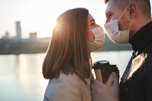 Young couple, a man and a woman in medical masks and gloves, drink coffee from disposable cups on the street and look at each other against the background of the river and the city. Quarantine.