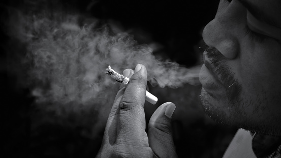 Focus at hand and close up man with mustache smoking cigarette and exhaling smoke in black and white with dark tone style, bad habit in unhealthy concept