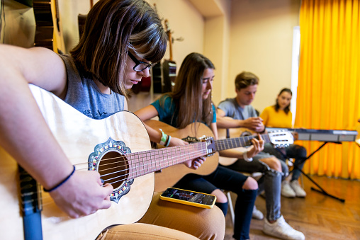 A group of students learns how to play musical instruments. The girl in the front is tuning her guitar with a mobile phone app