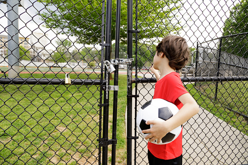 A little boy in a red jersey holding a soccer ball/ football stands by a locked fence.  He cannot get into the field to play.   You can see the field beyond the fence.  The fence is locked with a padlock and chain.