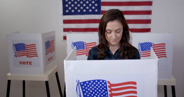 Front view young woman in booth at polling station MS front view Caucasian American woman in plaid shirt in voting booth, casting vote at polling station. US flag on wall in background polling place photos stock pictures, royalty-free photos & images