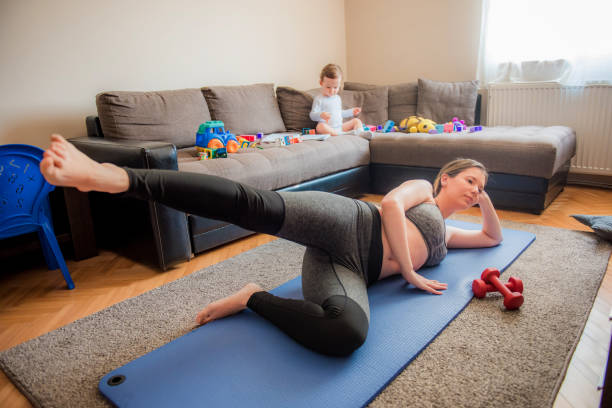 young mom exercising on a floor in a living room while toddler playing in a background - mother exercising baby dieting imagens e fotografias de stock