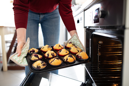 Close-up of woman taking out freshly baked blueberry muffins tray out of oven. Female holding a muffin pan with freshly baked blueberry muffins in kitchen.