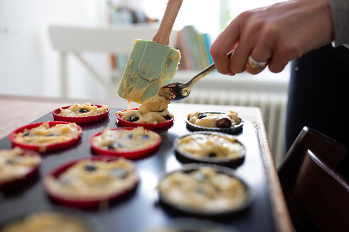 Close-up shot of woman scooping batter into muffin tin at the kitchen counter. Woman cooking blueberry muffins, pouring batter in baking dish on table.