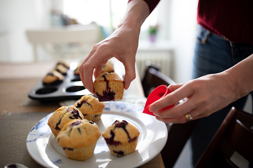 Close-up of a woman taking out blueberry muffins from paper mould in kitchen. Female serving freshly made in a plate.