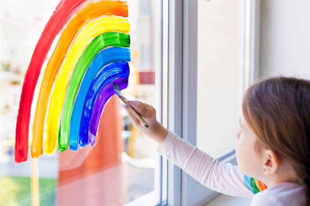 bambini a casa. una bambina dipinge un arcobaleno su una finestra durante la quarantena per la pandemia di coronavirus. flash mob sociale a sostegno della società. stiamo tutti bene. resta a casa - child multi colored painting art foto e immagini stock