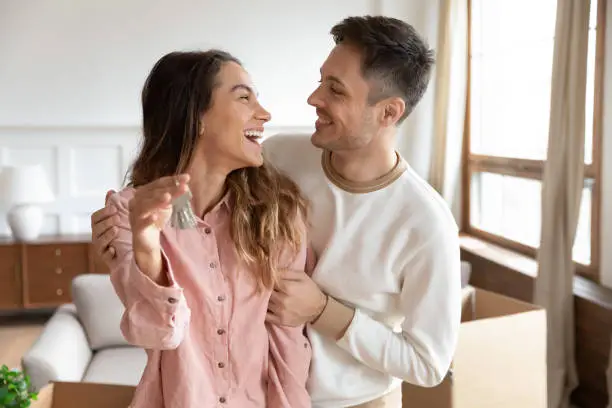 Joyful young man embracing smiling mixed race wife holding keys in hands. Excited family couple celebrating moving in new house apartment. Happy homeowners cuddling, amazed by real estate purchase.