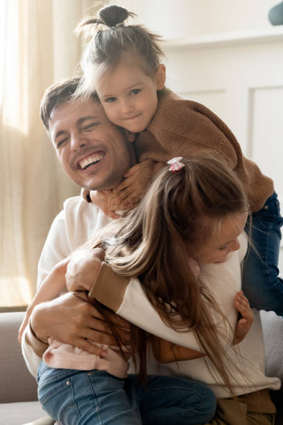 Playful little children embracing cuddling joyful father. Vertical image playful little children embracing cuddling joyful father, welcoming at home. Happy young daddy enjoying sweet tender moment, glad to see small kids siblings. having fun together. simple living photos stock pictures, royalty-free photos & images