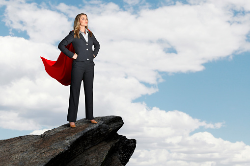 A businesswoman wearing a red cape strikes a confident pose and stands on a mountain peak as she looks at toward the cloud filled sky.