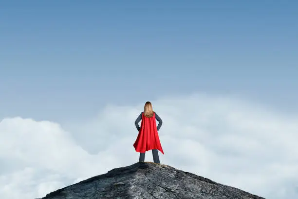 A businesswoman wearing a red cape strikes a confident pose and stands on a mountain peak as she looks at toward to blue sky and clouds below.