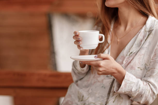 young woman on the balcony holding a cup of coffee or tea in the morning. she in hotel room looking at the nature in sumer. girl is dressed in stylish nightwear. relax time - espresso women cup drink imagens e fotografias de stock