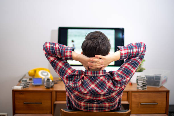 Person From Behind Looking at Monitor While Editing Videos Woman edits home movies in her home office during the coronavirus pandemic. She looks at the computer screen with her hands behind her head. The view is from behind. female film editor stock pictures, royalty-free photos & images