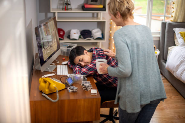 Mom Brings Coffee to Daughter Asleep on her Video Editing Job Woman edits home movies in her home office during the coronavirus pandemic. She is asleep with her head in her arms on her desk. Her mom brings in a cup of coffee to offer to her sleeping daughter. female film editor stock pictures, royalty-free photos & images