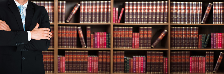 Midsection of lawyer with arms crossed standing against books in shelves