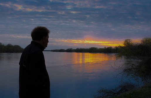 Older man watching sunset on Missouri River; Missouri, Midwest; setting sun reflects in water; woods at distance