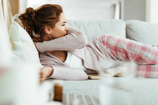 Sick woman sneezing into her elbow while lying down on sofa at home.