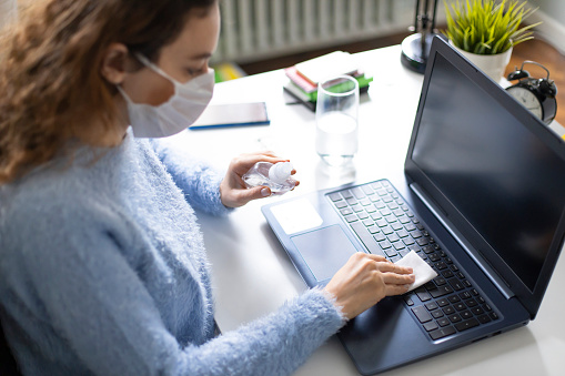 Young woman wiping laptop computer with disinfectant wipe while working from home