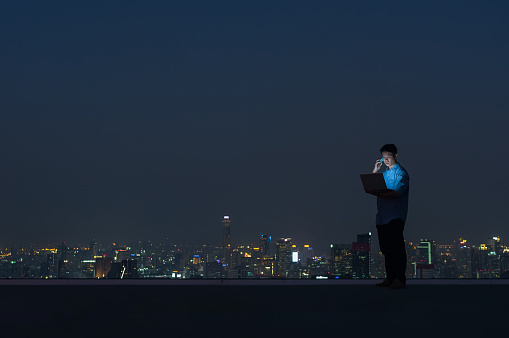 Asian businessmen stand at the top of the building. Use the laptop computer and smartphone with the scenery of the night view of the city, Business success and technology concept.