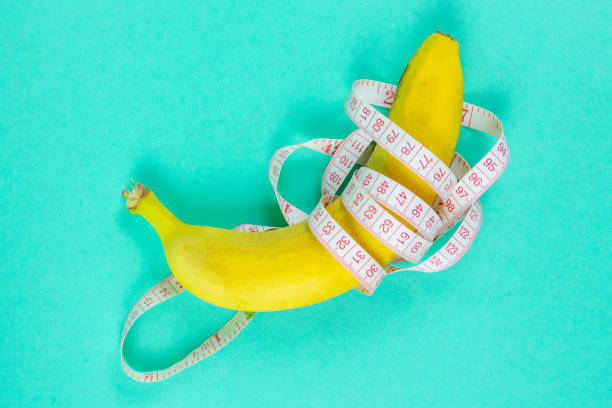 istock Banana with tape for measuring figure. Centimeter ruler spinned around fruit. Tape wrapped around banana isolated on mint background. Weight loss, healthy food and slim body concept 1218173431