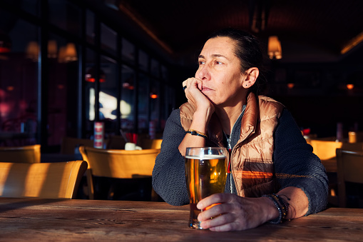 Unhappy, depressed and lonely middle age woman sitting at the bar with a beer glass.