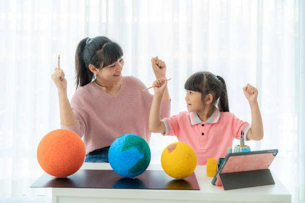 asian elementary schoolgirl with mother painting the moon in science class learning about the solar system via video conference with teacher and other classmates at home, homeschooling and distance learning. - earth globe mother child imagens e fotografias de stock