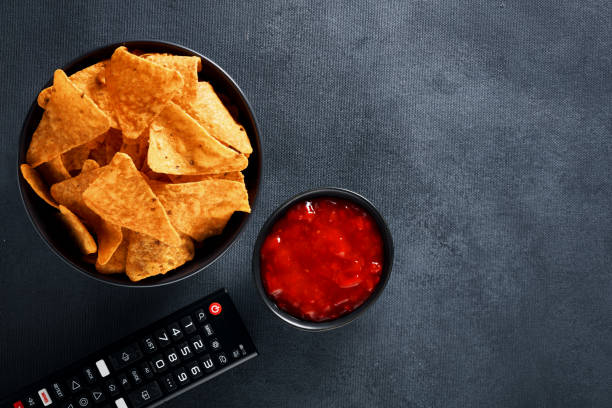 Mexican nachos in a black bowl with hot salsa dip sauce and tv remote on black background Mexican nachos in a black bowl with hot salsa dip sauce and tv remote on black background. Watching tv and eating snacks. Top view with copy space. remote control on table stock pictures, royalty-free photos & images