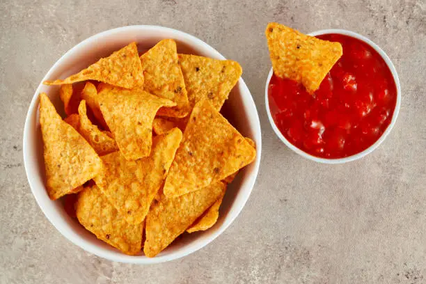 Mexican nachos or tortilla chips in a bowl with one nachos is dipped into chili salsa sauce. Macro top view.
