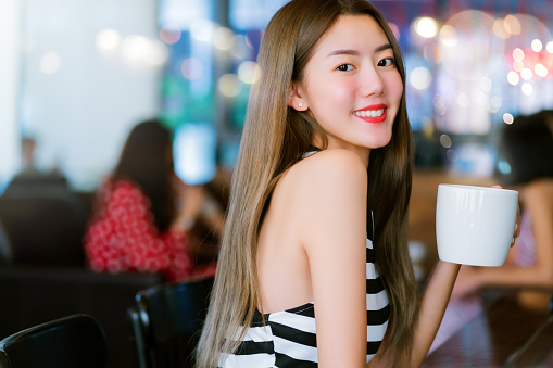 Young attractive beautiful asian woman big smile hand hold white coffee cup in coffee shop cafe restaurant tone color blur background