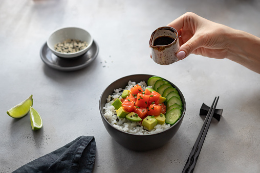 womans hand holds a sauceboat with soy sauce over a dark bowl with salmon, rice, avocado and cucumber. traditional Hawaiian raw fish salad. horizontal image, gray concrete background.