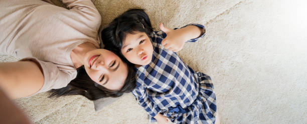 felice famiglia asiatica madre e figlia facendo una divertente foto selfie viso mentre si sdraiano sull'uso del pavimento nello sfondo della casa del soggiorno. - floor two parent family couple home interior foto e immagini stock