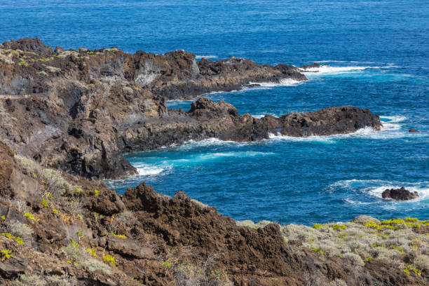 formazione rocciosa vulcanica, scogliere di lava nera sulla costa rocciosa con onde bianche schiacciante sull'oceano atlantico. sfondo cielo blu. la palma, isole canarie. - rocky coastline spain la palma canary islands foto e immagini stock