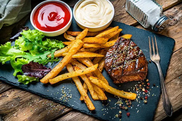 Grilled tenderloin with French fries and salad served on a slate board. Two bowls with ketchup and mayonnaise are at the top of an horizontal frame. A salt shaker complete the composition. High angle view. High resolution 42Mp studio digital capture taken with SONY A7rII and Zeiss Batis 40mm F2.0 CF lens