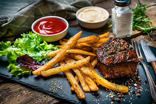 Grilled tenderloin with French fries and salad served on a slate board. Two bowls with ketchup and mayonnaise are at the top of an horizontal frame. A salt shaker complete the composition. High resolution 42Mp studio digital capture taken with SONY A7rII and Zeiss Batis 40mm F2.0 CF lens