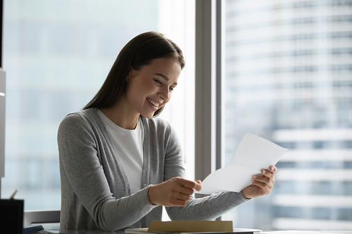 Smiling millennial girl sit at desk in office reading good news in postal letter, happy young woman consider paperwork post correspondence, receive positive reply answer or promotion notice