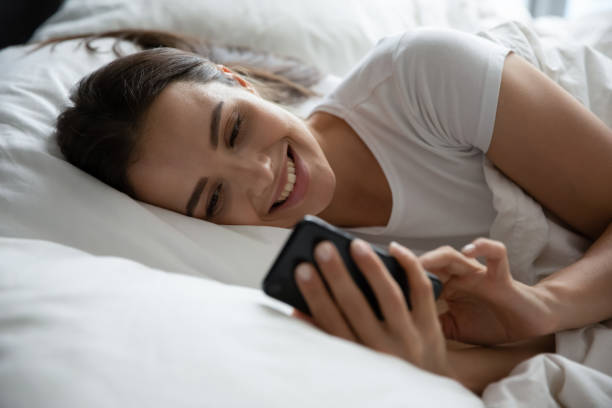 istock Smiling young woman relaxing in bed using cell 1218204753